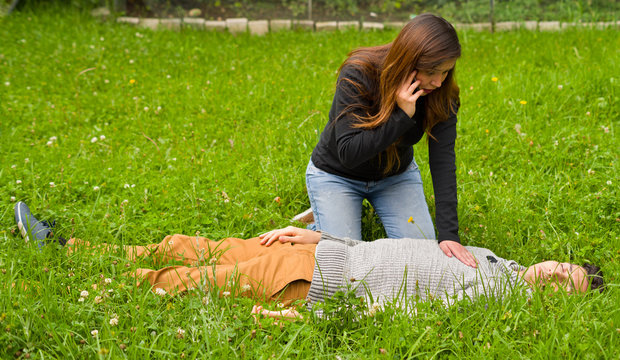 Beautiful woman giving first aid to a handsome young man and using her cellphone to call the ambulance, cardiopulmonary resuscitation, in a grass background