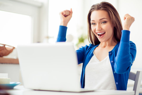 Oh yeah! Happy amazed young girl is celebrating. She is excited and with open mouth, wearing casual clothes, gesturing, sitting in light designed cafe in front of laptop