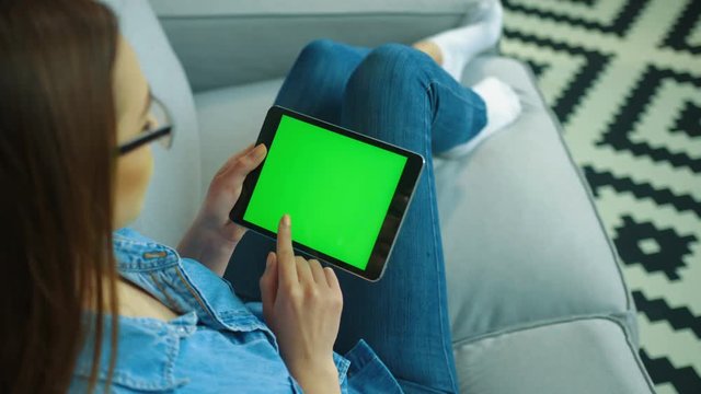 Young woman using black tablet computer with green screen while sitting on the couch in the living room. Woman hands scrolling, zooming pages. Chroma key. top view