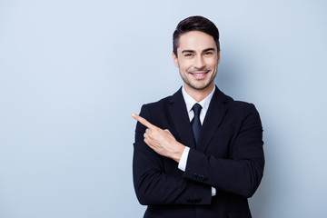 Close up portrait of young successful brunete stock-market broker guy on the pure light blue background, he is smiling, wearing suit with tie and is pointing on a copyspace with his finger