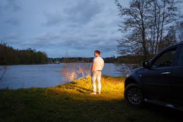 A young man stands in the light of the headlights of the cars on the background of beautiful scenery next to the road. A traveler admires the sunset.