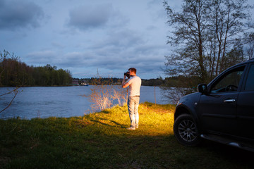 A young man stands in the light of the headlights of the cars on the background of beautiful scenery next to the road. A traveler admires the sunset. Man taking picture of sunrise car travel.