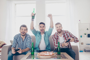 Yes! Team of winners! Happy joyful men are sitting on sofa and playing video games with beer and pizza, smiling, gesturing, enjoying