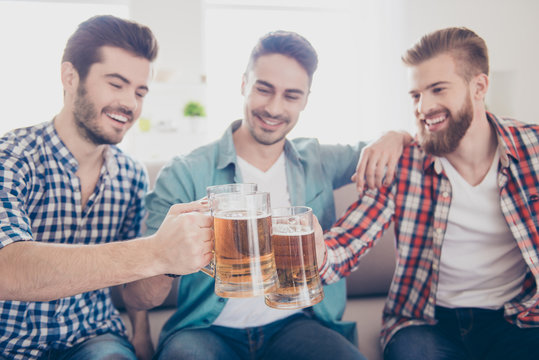Close up portrait of three happy men friends, clinking with glasses of beer, smiling, alll are in casual shirts and jeans, sitting on couch at home