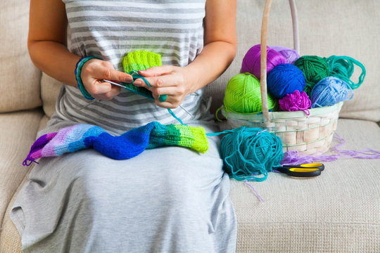 A woman in a gray dress knit socks from colored yarn. Balls of colored yarn are in the basket.