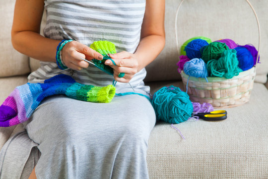 A woman in a gray dress knit socks from colored yarn. Balls of colored yarn are in the basket.