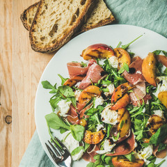 Arugula, prosciutto, mozzarella and grilled peach salad in white plate over blue napkin and wooden board, top view, square crop