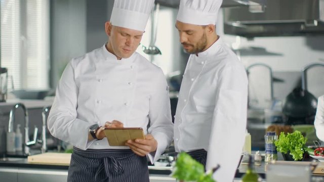 Two Famous Chefs Exchange Ideas About Video Blog Recipe they Watching on Tablet. They are Working in Big Restaurant Kitchen. Shot on RED EPIC-W 8K Helium Cinema Camera.