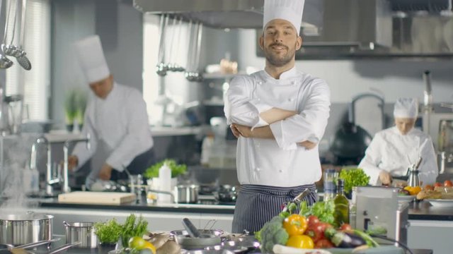 Famous Chef of a Big Restaurant Crosses Arms and Smiles in a Modern Kitchen. His Staff in Working in the Background. Shot on RED EPIC-W 8K Helium Cinema Camera.