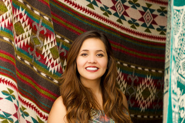 Close up of a beautiful smiling young woman posing for camera in front of andean traditional clothing, colorful fabrics background