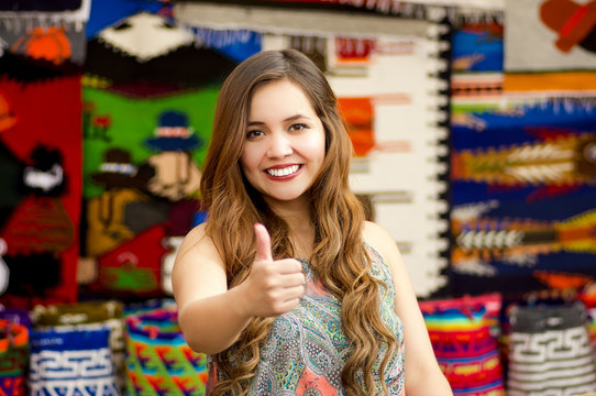Beautiful smiling young woman with humbs up and posing for camera, with colorful andean traditional clothing fabrics background