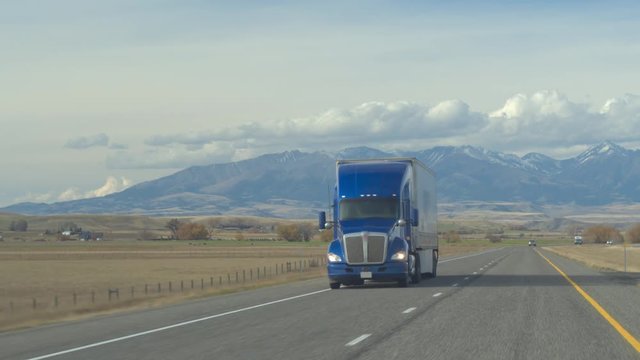 CLOSE UP: Freight container semi truck transporting goods driving along the scenic rural highway past pasture fields and farmlands. Lorry trucking load, snowy Rocky Mountain Range in the backgrounds