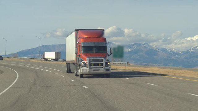 CLOSE UP: Freight container semi truck hauling goods, cars, SUVs and pickups traveling along the busy traffic highway running through prairie countryside with snowy mountain-peaks in the background