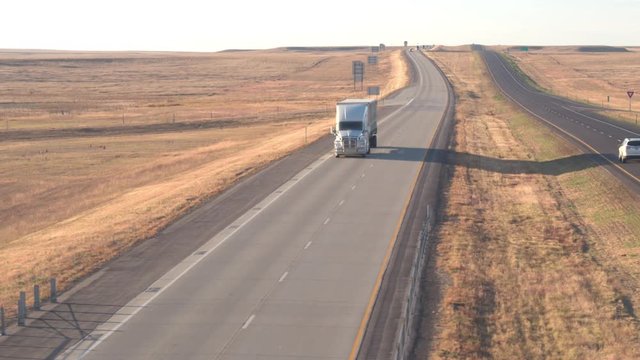 AERIAL CLOSE UP: Flying above freight semi truck transporting goods, cars on road trip, people in SUVs traveling along the busy rural highway running through vast grassy fields in American countryside
