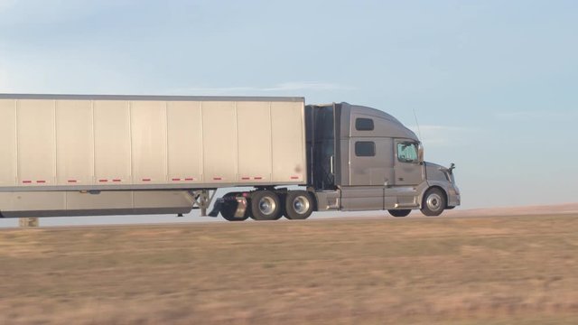 AERIAL, CLOSE UP Freight container semi truck hauling goods driving along the empty interstate highway through the prairie fields in the Great Plains, America. Trailer shipping cargo on sunny morning