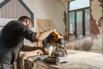 A young male construction carpenter saws a modern circular saw with a wooden board in the workshop room, a wooden sawdust fly to the sides, a rear view sideways - obrazy, fototapety, plakaty