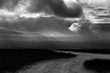A mountain road with some moody clouds in the sky and faint sunrays in the background