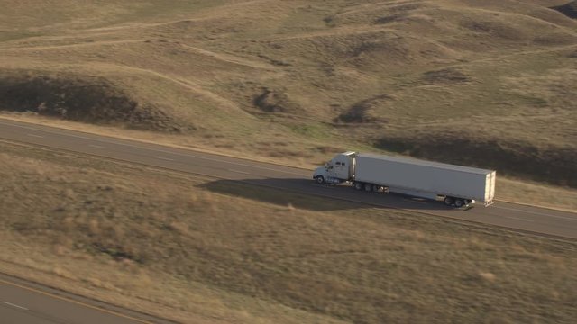 AERIAL Flying above freight container semi truck driving along the empty highway running through sunny bumpy hilly landscape rolling into the distance. Trailer transporting goods across United States