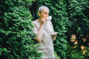 Portrait of positive middle aged woman with short gray hair drinking and holding white cup of tea...