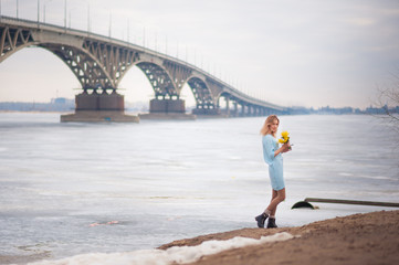 A beautiful girl in an amazing blue dress with mimosas on the background of a bridge over the river, she admires, dances, graces and mimosas, jumps from happiness