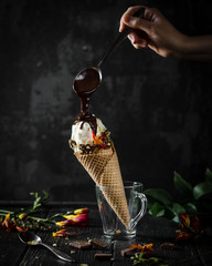 ice cream cone in a glass cup with woman's hand holding a spoon of melted chocolate pouring on ice...