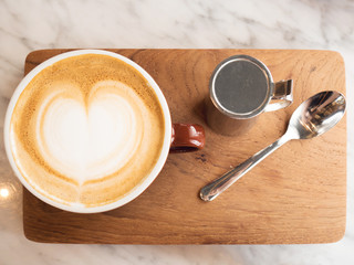top view of a cup of hot coffee with heart latte art and milk mug and spoon on wood plate with marble table as background