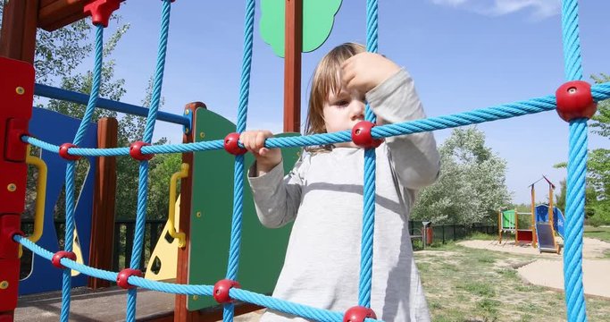 three years old blonde child playing climbing in ropes ladder in outdoor playground, in public Park Valdebebas, Madrid Spain
