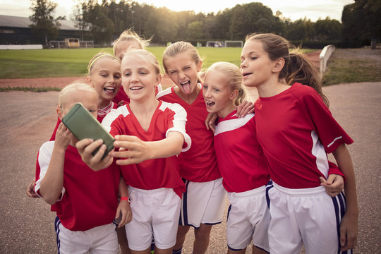 Cheerful soccer girls taking selfie on footpath against field