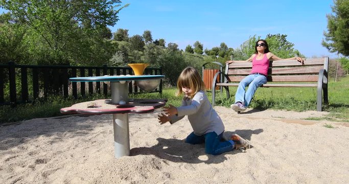 three years old blonde child playing with sand in playground next to mother sitting in bench resting and watching
