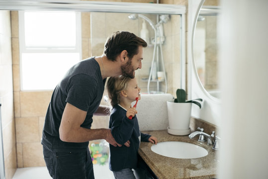 Side view of father watching daughter brushing teeth at sink in bathroom