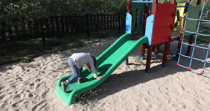 side view of three years old blonde child playing trying to climb on green plastic small slide and falling. Outdoor playground, in public Park Valdebebas, Madrid Spain
