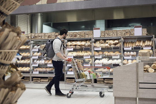 Side view of man walking with cart while looking at breads in supermarket