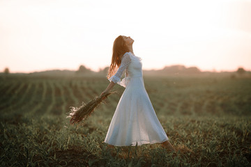 Young woman in long white lace dress on cornfield. Side view.