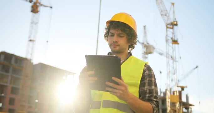 Concentrated young builder in yellow helmet and green vest typing on tablet for work on unfinished construction background.