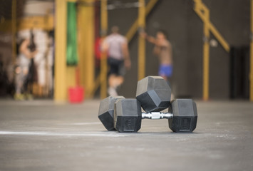 two dumbells sitting in the middle of a gym with out of focus athletes working out exercising in the background 