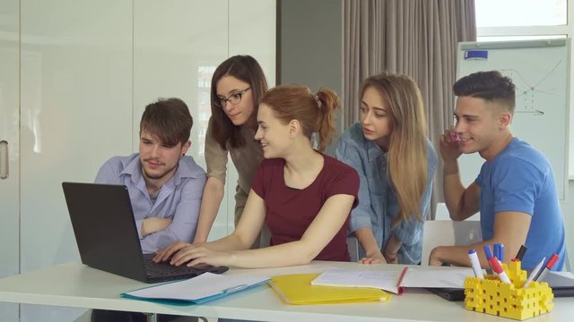Five creative young people showing teamwork at the office. Handsome caucasian guys sitting at the table from the both sides of three girls. Two pretty women giving some advices to their female