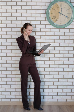 Smiling Businesswoman Working On Laptop And Talking On Smart Phone In A Bright Office On The Background Of A White Brick Wall. Looking Away From The Camera. Girl In Elegant Office Suit.