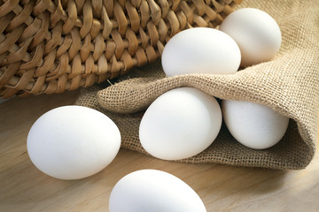 White chicken eggs wrapped in burlap next to a basket on a wooden table