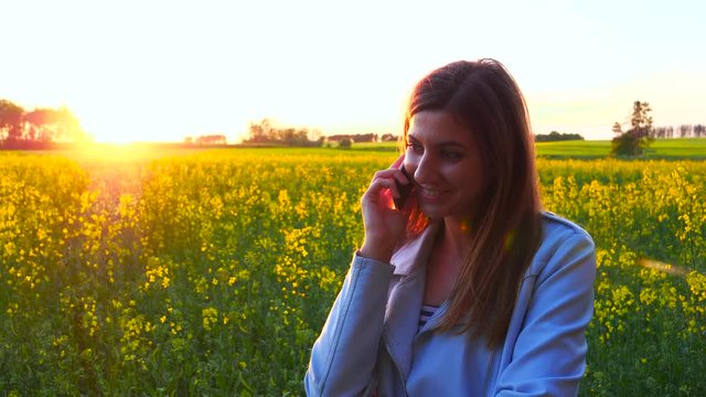 Young beautiful woman using smartphone on the silhouette of the sunset field.