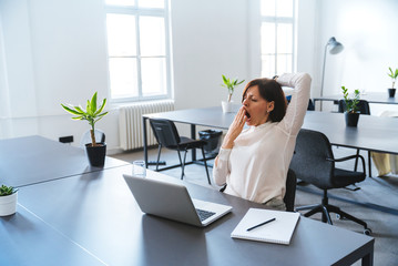 Young business woman yawning at a modern office desk in front of laptop.