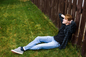 Handsome young man sitting on the grass near the brown wooden fence