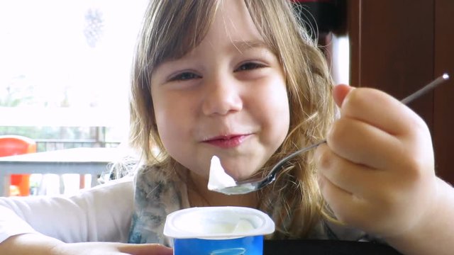 portrait of funny blonde caucasian baby three years old with white shirt, sitting indoor, eating yogurt with spoon, looking, speaking and making fun 
