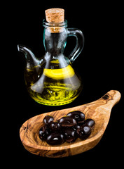 Black olives in wooden spoon and bottle of virgin olive oil on black background