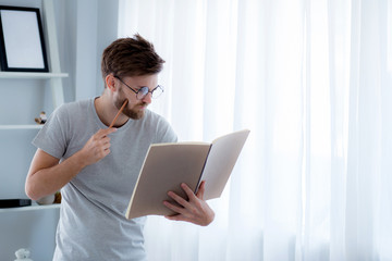 Handsome guy in eyeglasses is reading book preparing exam with standing at the living room, education concept