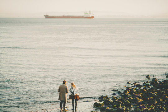 Silhouettes of young couple: man in coat and woman in skirt and jeans jacket standing on sand near water of winter beach in Lisbon with huge cargo barge in distance and hills on misty horizon
