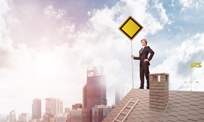 Young businessman on house brick roof holding yellow signboard and looking at city. Mixed media