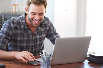 Happy young adult caucasian man smiling joyfully while looking at his phone, reading his latest messages to catch up with his social media as a way of procrastinating.