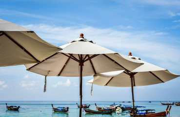 White beach umbrellas against light blue sky with soft clouds. Tourist boats floating near the beach in the  background.