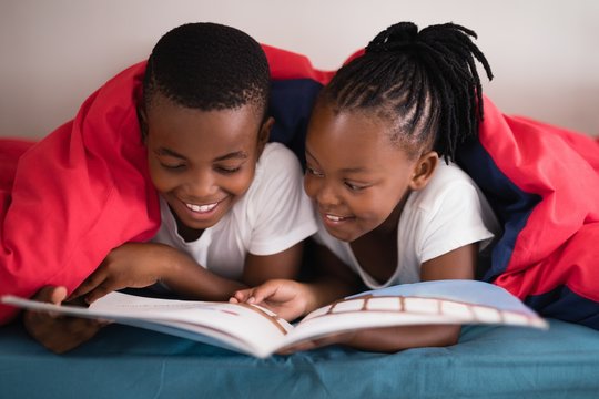 Smiling Siblings Reading Book Together While Lying On Bed