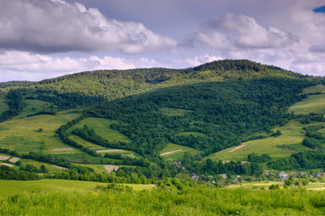 Obraz premium Scenic landscape of the countryside near alpine mountains. View of green hills covered by trees. Summer natural background. Sunlight shines through the trees.